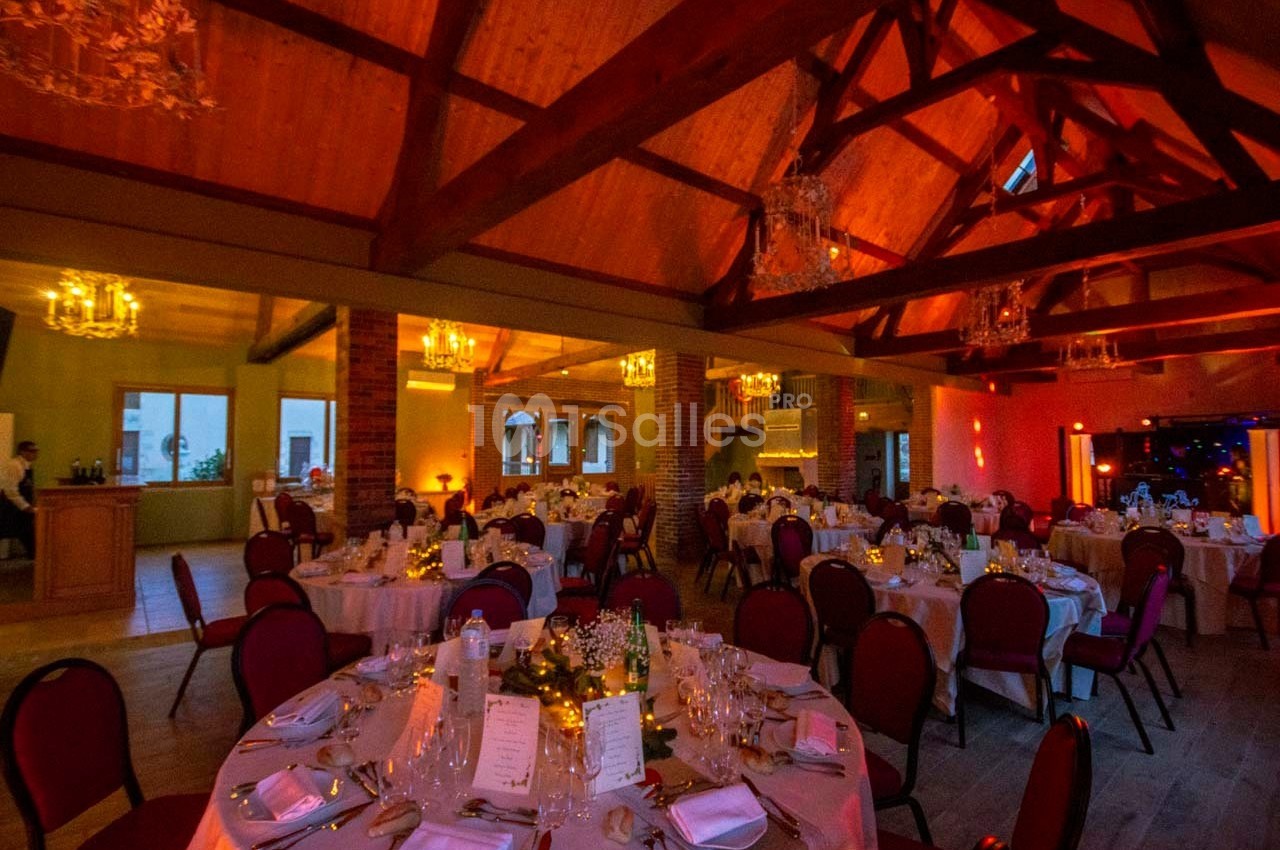 Salle de réception chaleureuse avec tables rondes dressées, éclairage tamisé et poutres apparentes en bois.