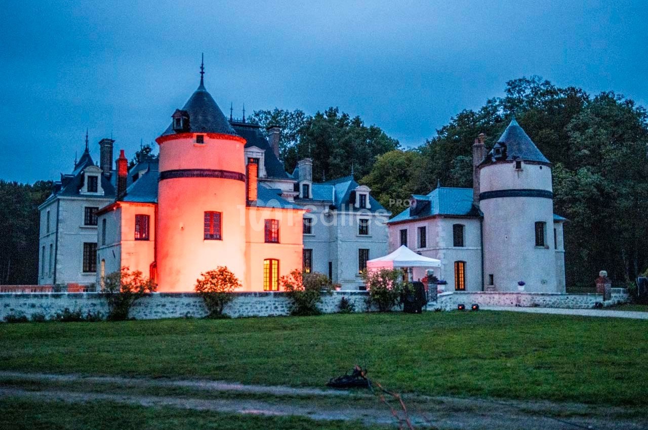 Château éclairé par des lumières rouges au crépuscule, entouré de verdure et d'arbres en arrière-plan.