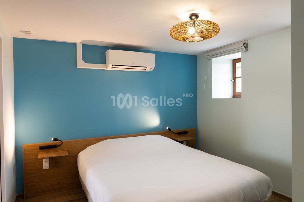 Chambre avec lit double, mur bleu, éclairage suspendu, climatisation et petite fenêtre avec store.