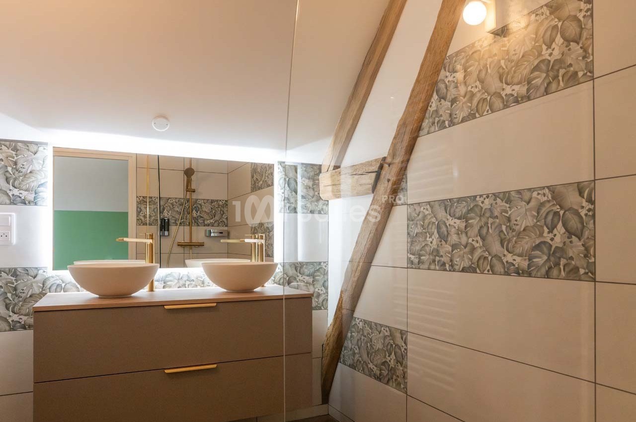 Salle de bain avec deux vasques blanches, miroir éclairé, poutre apparente et carrelage décoratif floral.