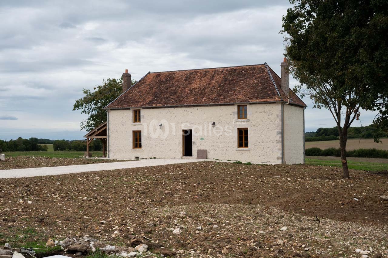 Maison en pierre blanche avec toit en tuiles rouges, entourée de terrain dégagé et d'arbres sous un ciel nuageux.