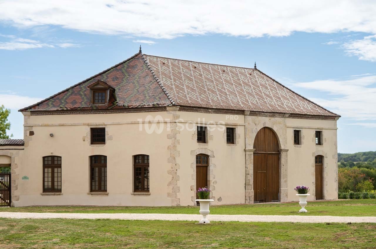 Bâtiment ancien avec toit en tuiles, grandes portes en bois et fenêtres, entouré de pelouse et de pots de fleurs.