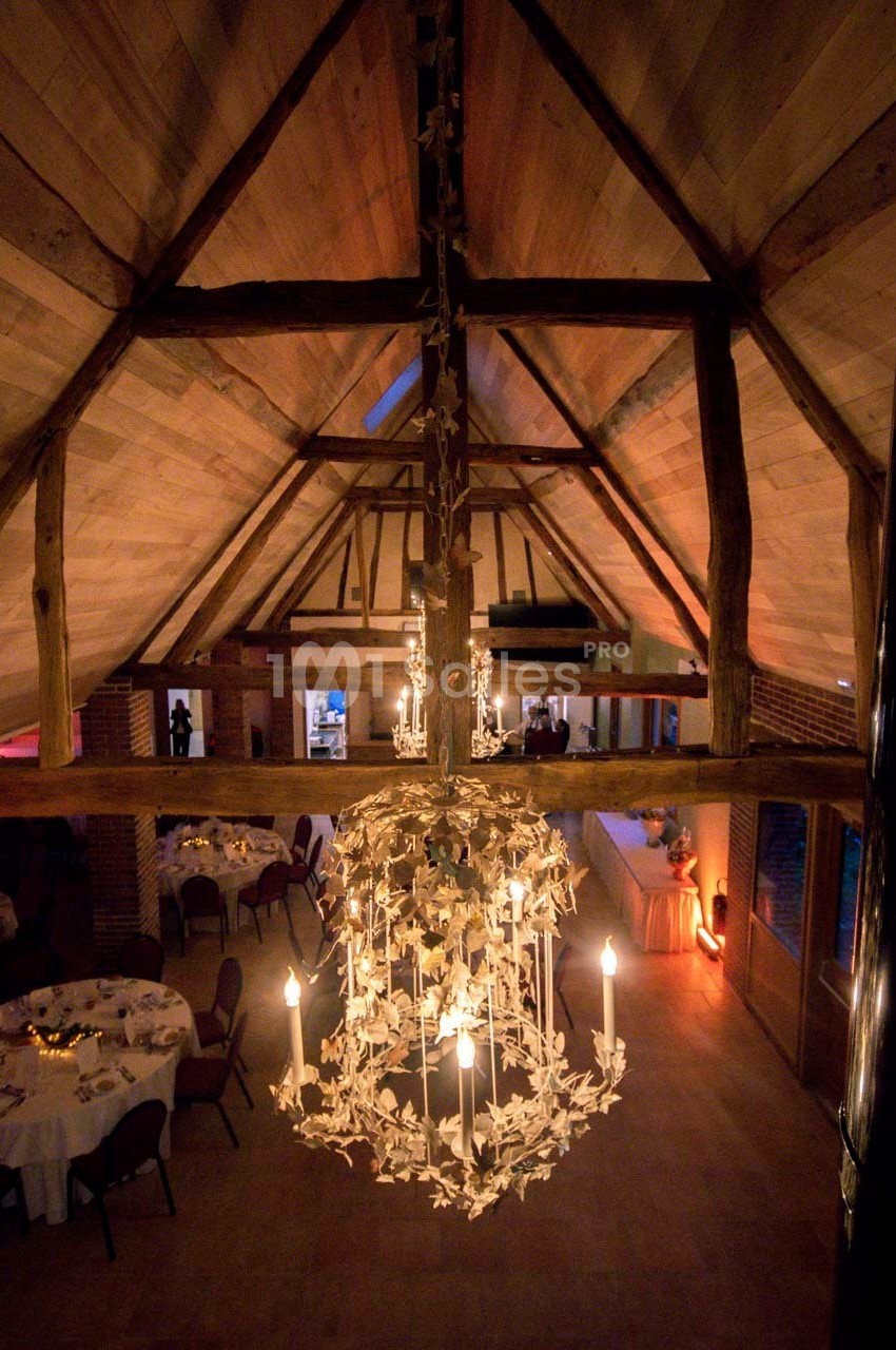 Vue intérieure d'une salle avec charpente en bois apparente, lustre décoratif et tables dressées pour un événement.