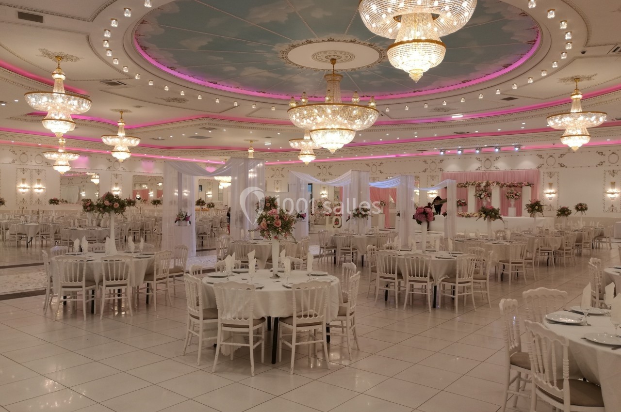 Salle de réception élégante avec tables dressées, lustres imposants et décoration lumineuse aux tons roses et blancs.