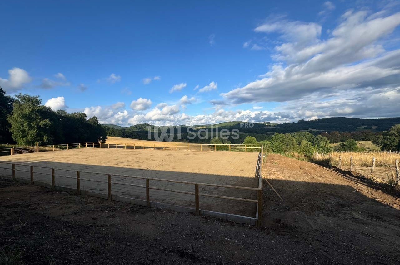 Vue d'une carrière équestre en sable entourée de barrières, avec un paysage vallonné et un ciel partiellement nuageux.