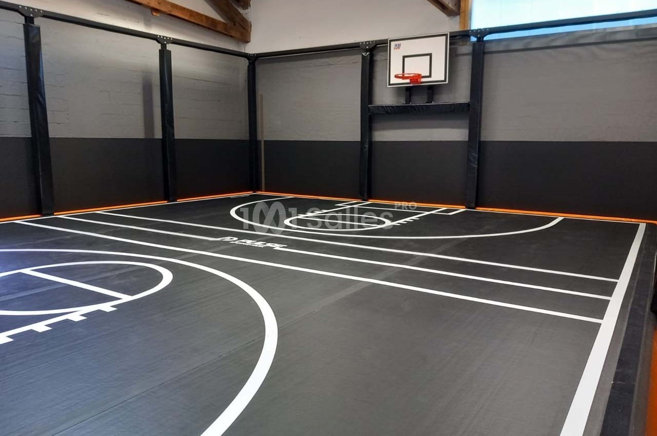 Terrain de basketball intérieur avec sol noir, lignes blanches et panneau de basket fixé sur un mur.