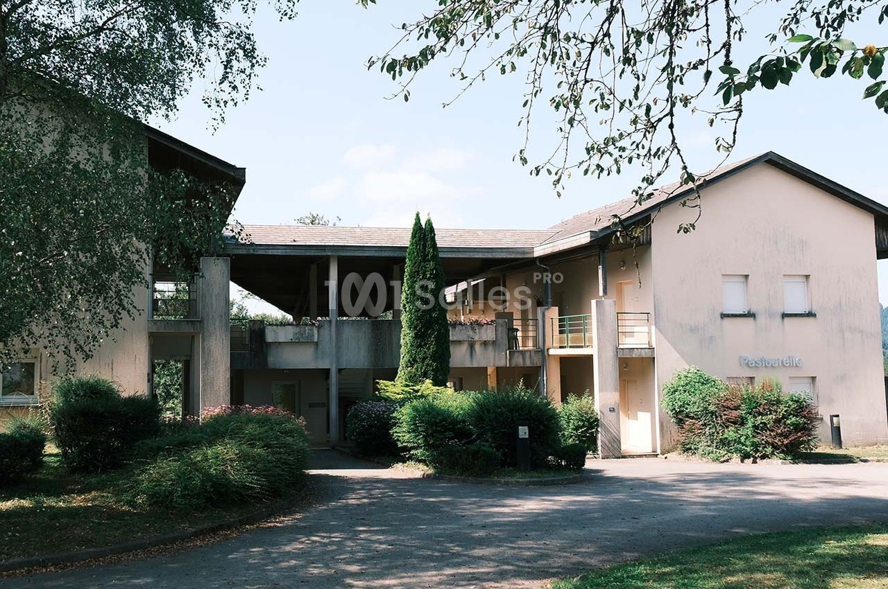 Bâtiment résidentiel beige entouré de végétation, avec un chemin d'accès et un arbre au premier plan.