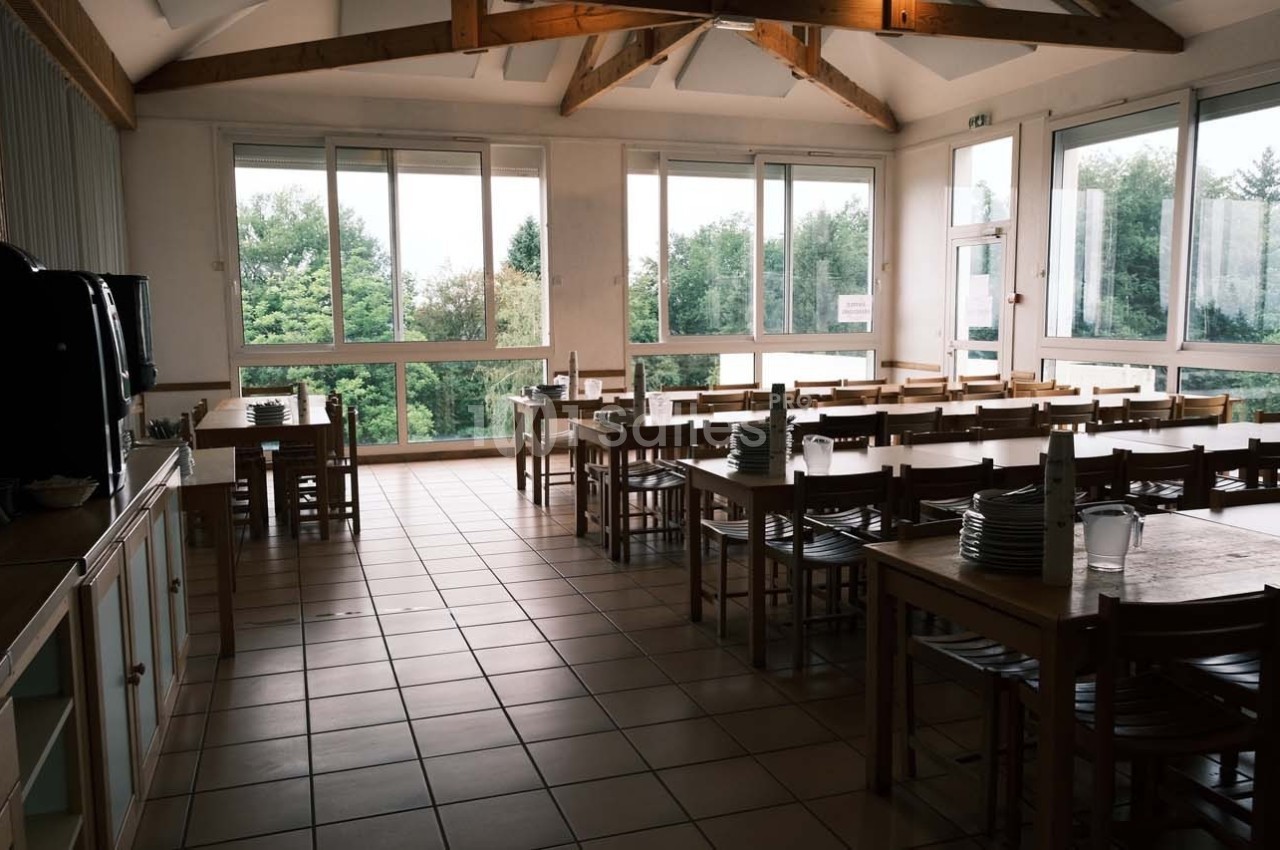 Salle à manger lumineuse avec de grandes fenêtres, des tables en bois alignées et de la vaisselle empilée.