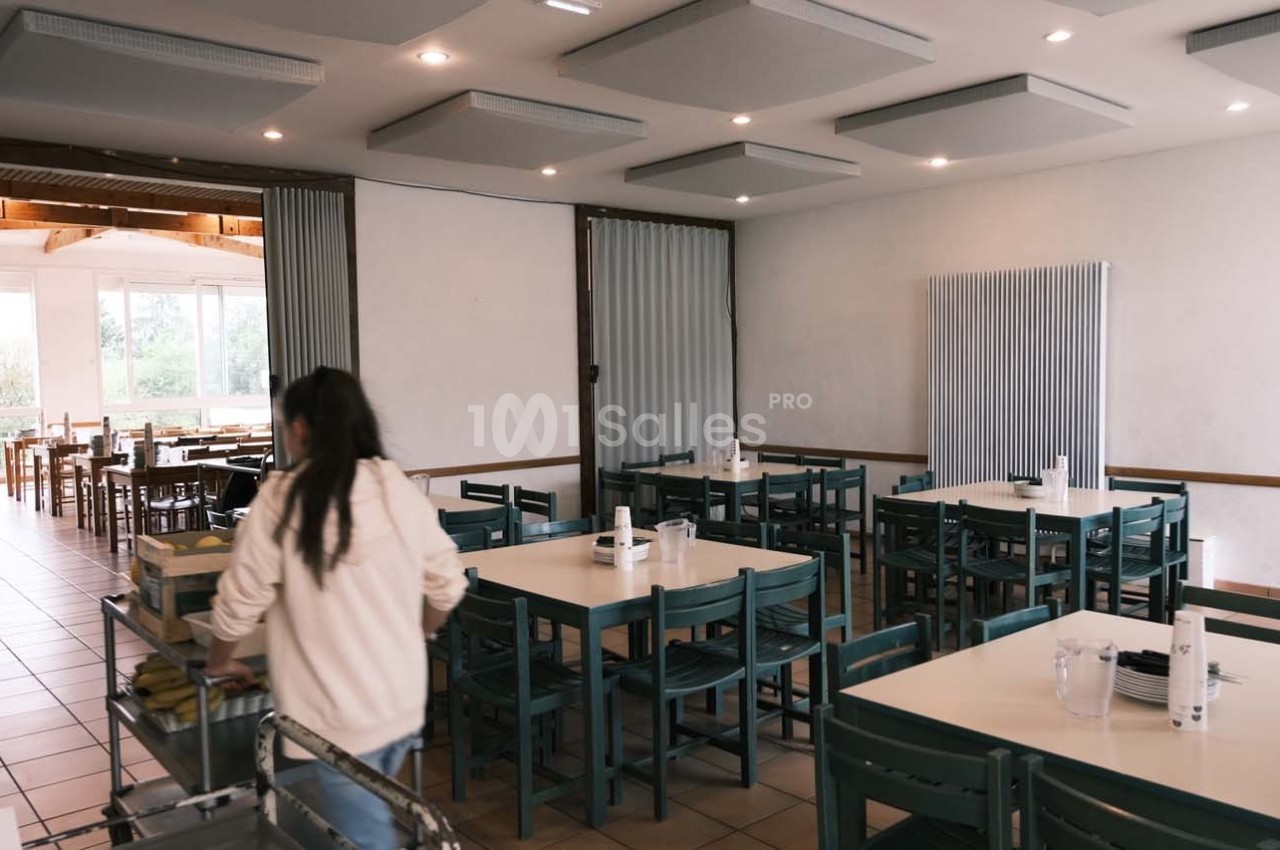 Une personne de dos range un chariot dans une salle de restaurant lumineuse avec des tables et des chaises alignées.