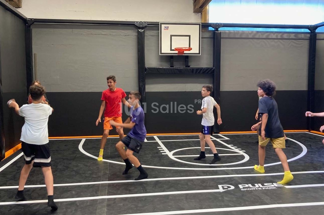 Des enfants jouent au basket dans un gymnase intérieur avec un terrain délimité et un panier mural.