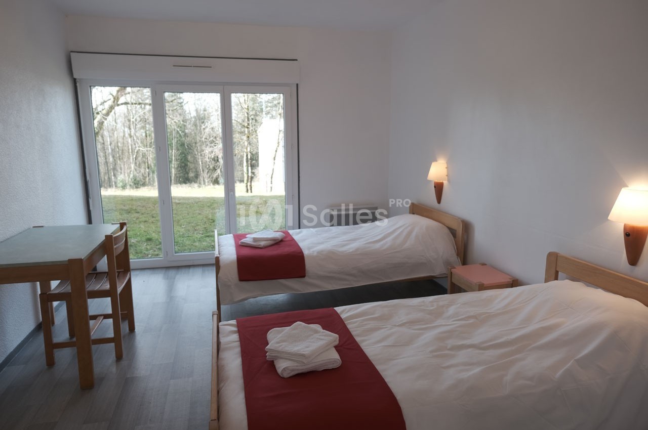 Chambre lumineuse avec deux lits simples, table en bois, et grande baie vitrée donnant sur un espace vert.