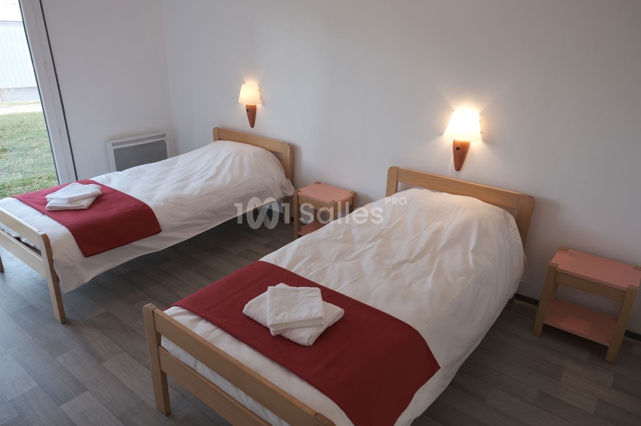Deux lits simples avec draps blancs et chemins de lit rouges dans une chambre lumineuse au sol en parquet.
