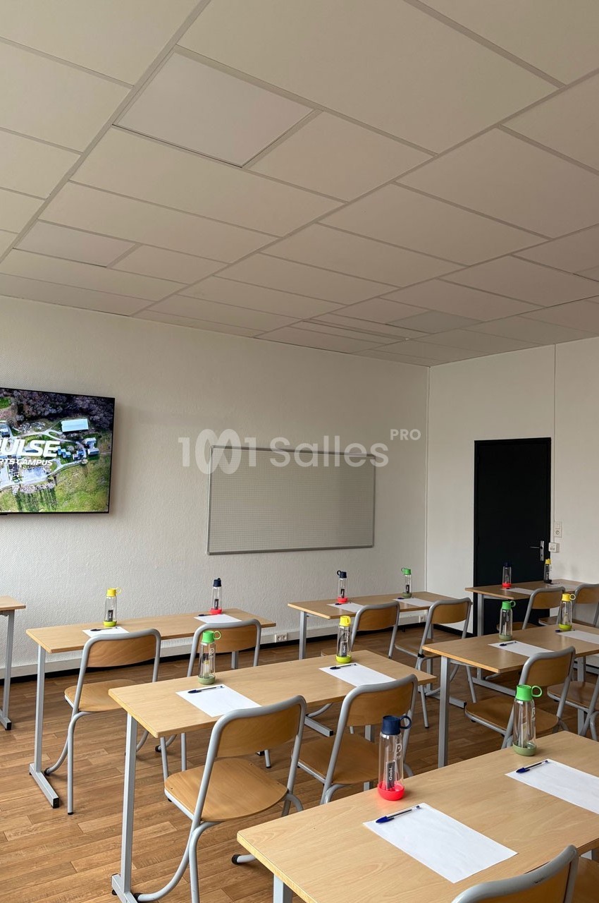 Salle de classe lumineuse avec des bureaux alignés, des bouteilles d'eau et un écran allumé au mur.