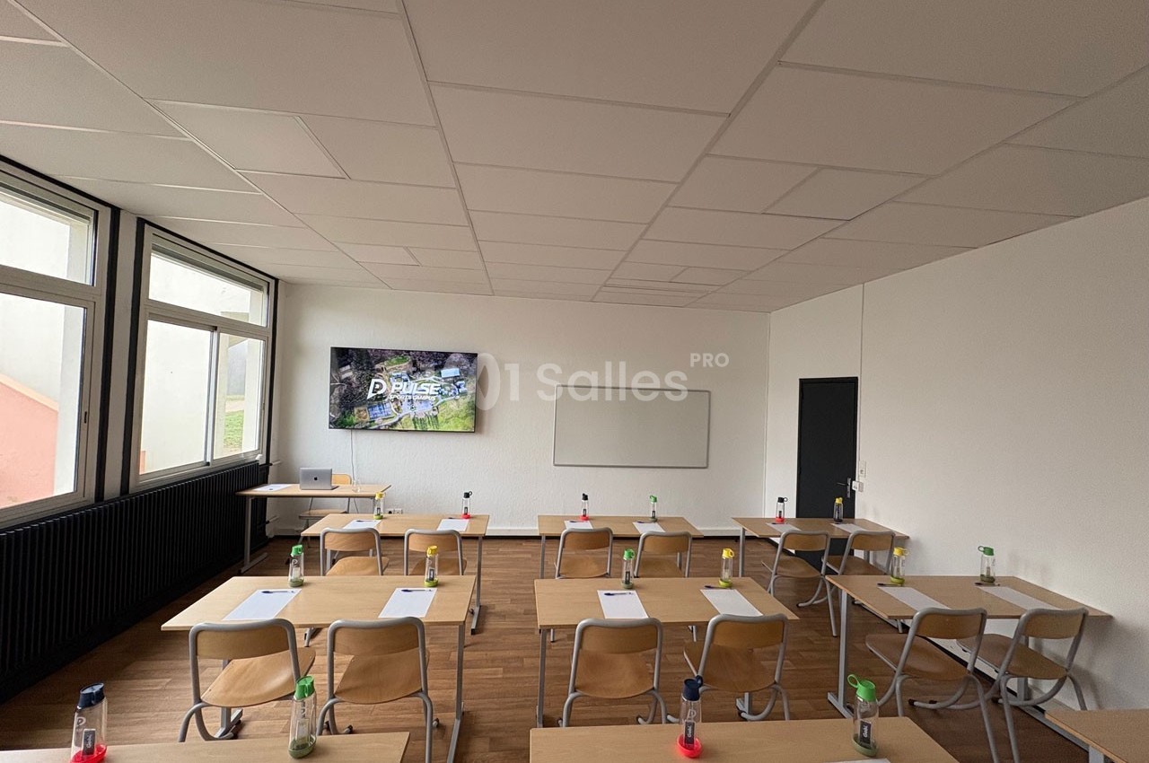 Salle de classe lumineuse avec des tables en bois, des chaises, un tableau blanc et un écran affichant une présentation.