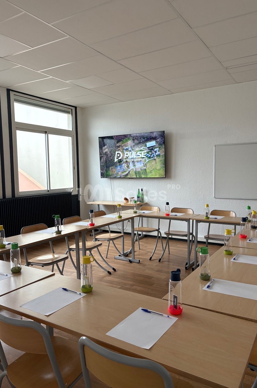 Salle de réunion lumineuse avec tables disposées en U, chaises en bois, écran mural et fournitures de bureau.