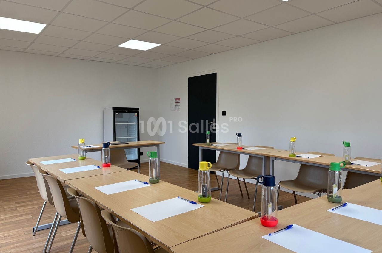 Salle de réunion lumineuse avec des tables disposées en U, des chaises, des bouteilles d'eau et des blocs-notes.