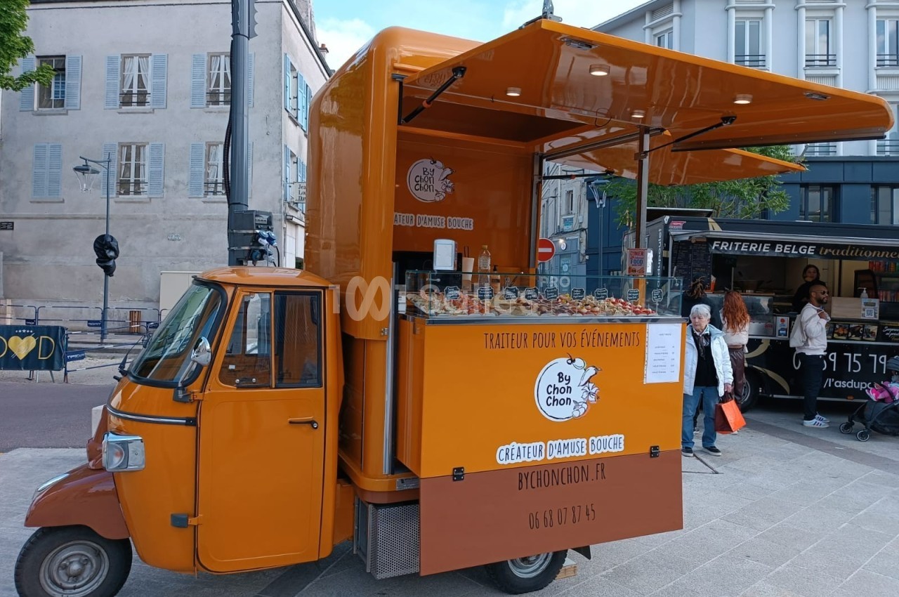 Triporteur orange aménagé en stand de traiteur, stationné sur une place urbaine avec des passants en arrière-plan.
