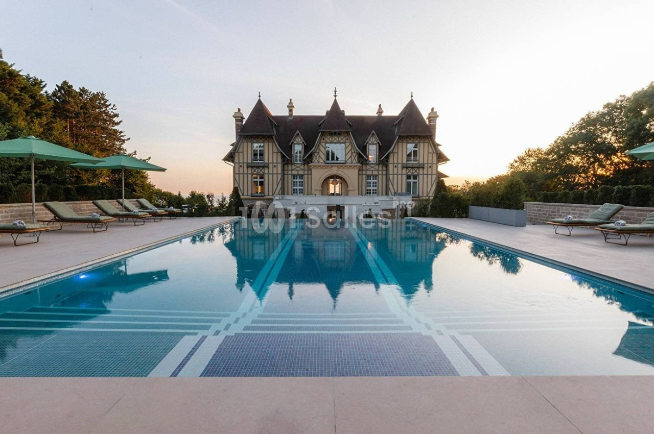 Piscine extérieure avec transats et parasols, devant un manoir à colombages entouré de verdure au coucher du soleil.
