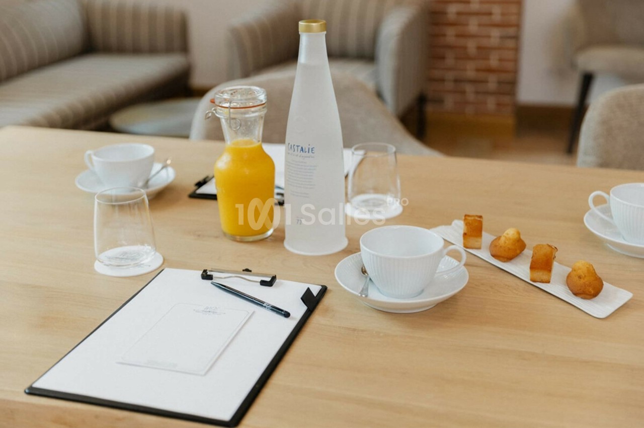 Table en bois avec carafe de jus d'orange, bouteille d'eau, tasses, verres, bloc-notes et assiette de petits gâteaux.