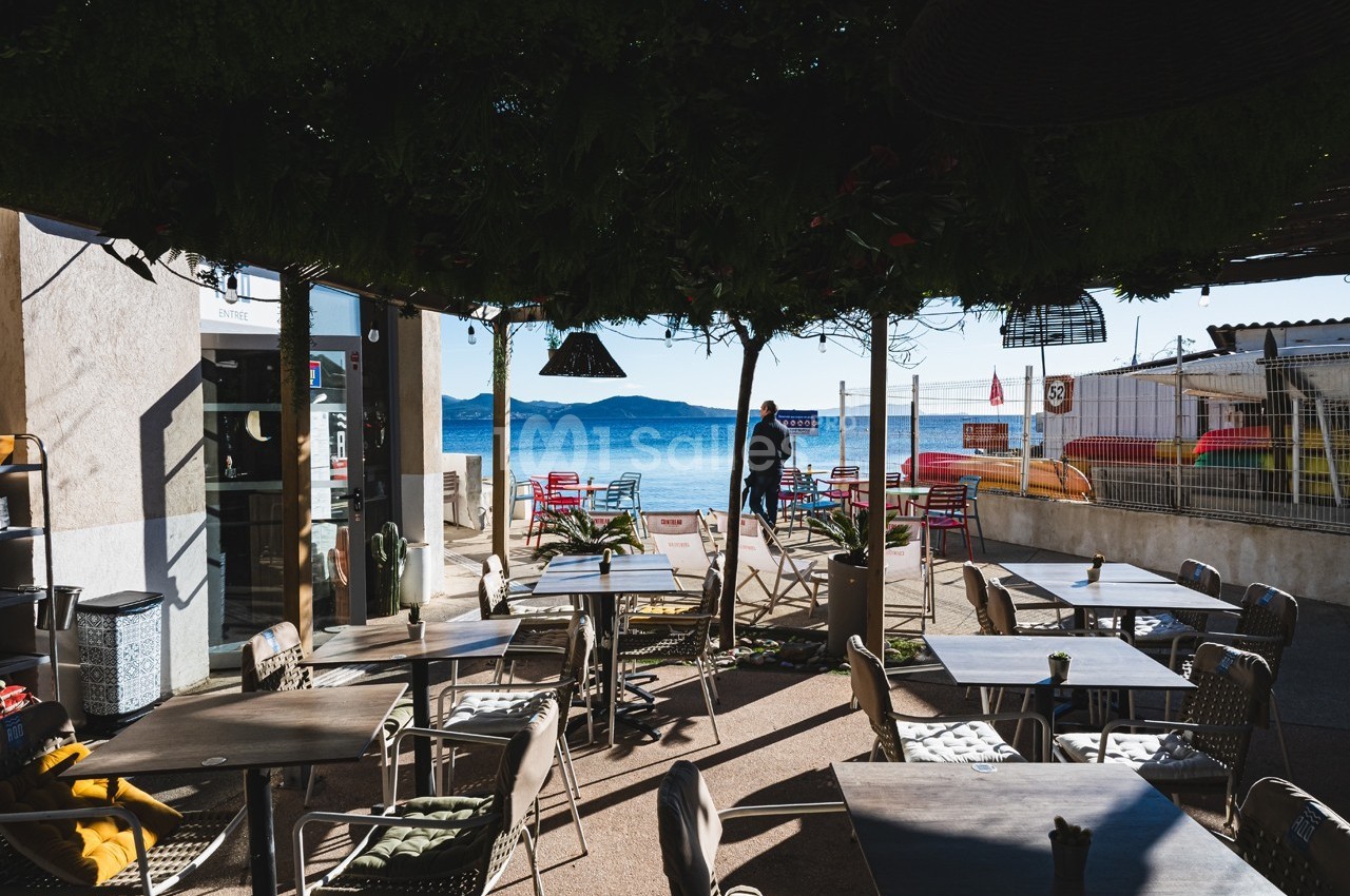 Terrasse ombragée d'un café avec vue sur la mer, tables et chaises disposées face au paysage côtier.