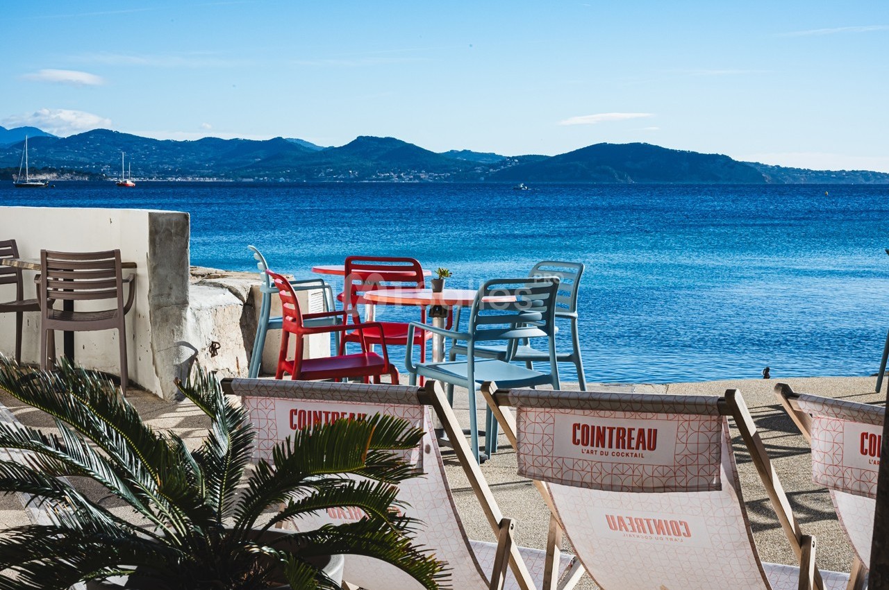 Terrasse en bord de mer avec des chaises colorées, des transats et une vue sur des montagnes à l'horizon.