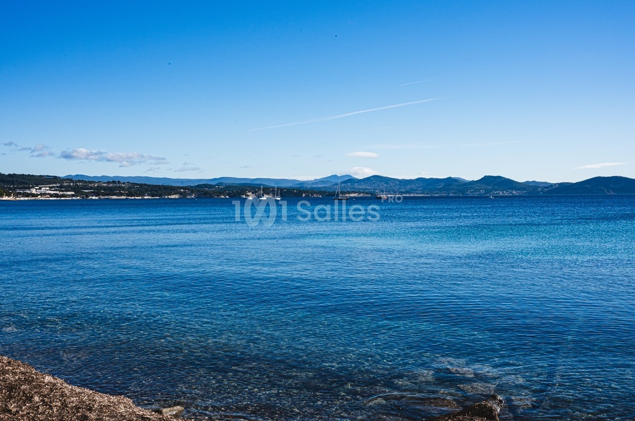 Vue sur une mer calme avec un rivage rocheux au premier plan et des montagnes à l'horizon sous un ciel dégagé.