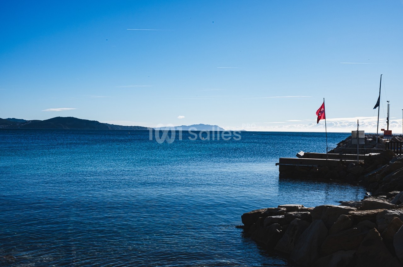 Vue d'un littoral avec une mer calme, un quai, des drapeaux et des collines à l'horizon sous un ciel dégagé.