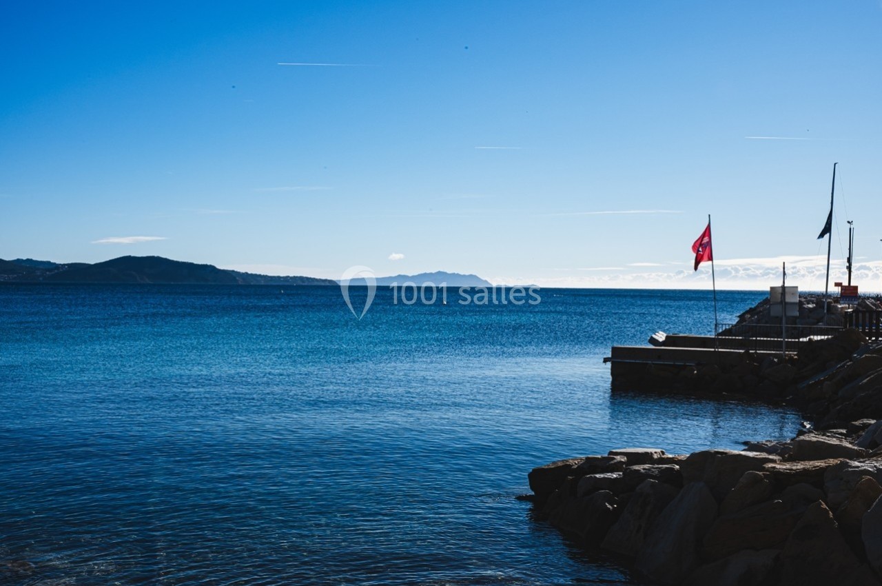 Vue d'un littoral avec une mer calme, un quai, des drapeaux et des collines à l'horizon sous un ciel dégagé.