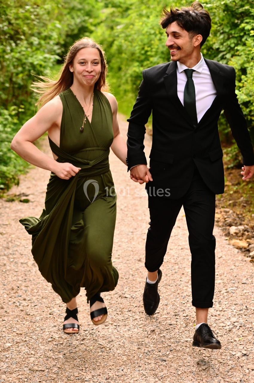 Un couple souriant court main dans la main sur un chemin entouré de verdure.