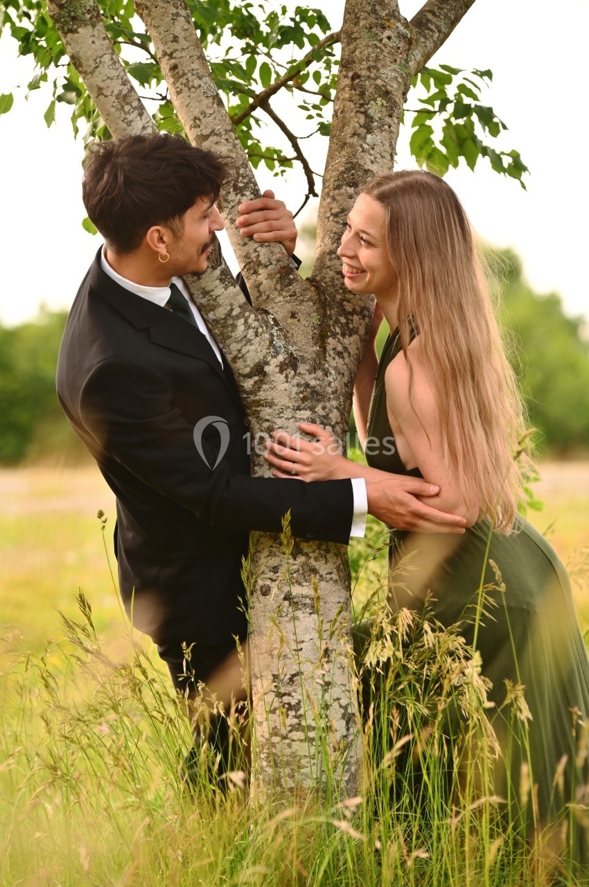 Un homme en costume et une femme en robe verte sourient en s'enlaçant autour d'un arbre dans un champ.