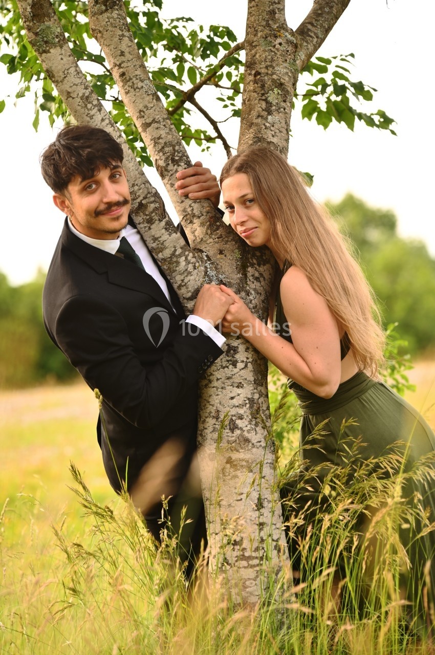 Un homme en costume et une femme en robe verte posent souriants de part et d'autre d'un arbre dans un champ verdoyant.