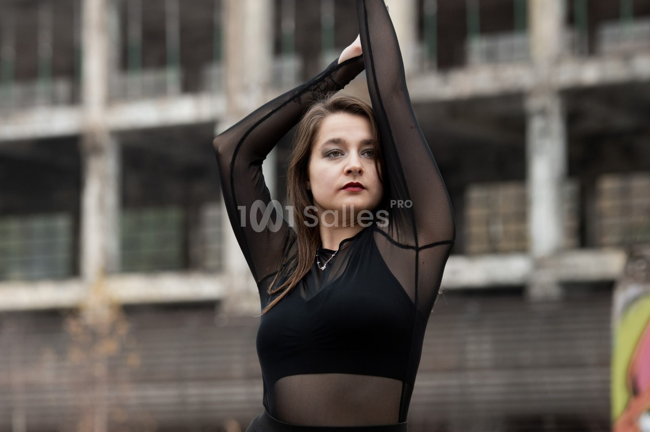 Femme en tenue noire transparente posant devant un bâtiment industriel délabré.