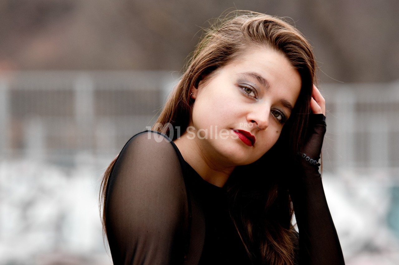 Femme aux cheveux longs bruns, portant un haut noir transparent, posant devant un fond flou extérieur.