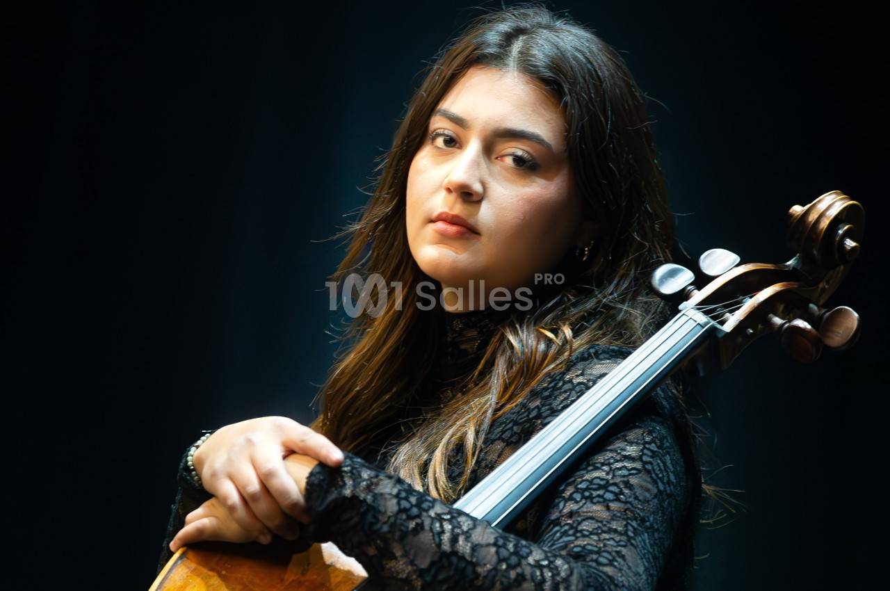 Une femme en tenue noire tient un violoncelle, éclairée par une lumière douce sur un fond sombre.