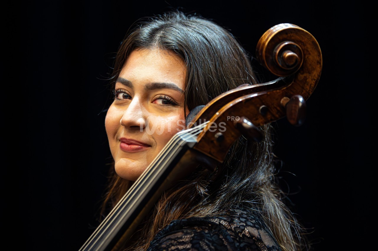 Une femme souriante tient un violoncelle devant un fond noir.