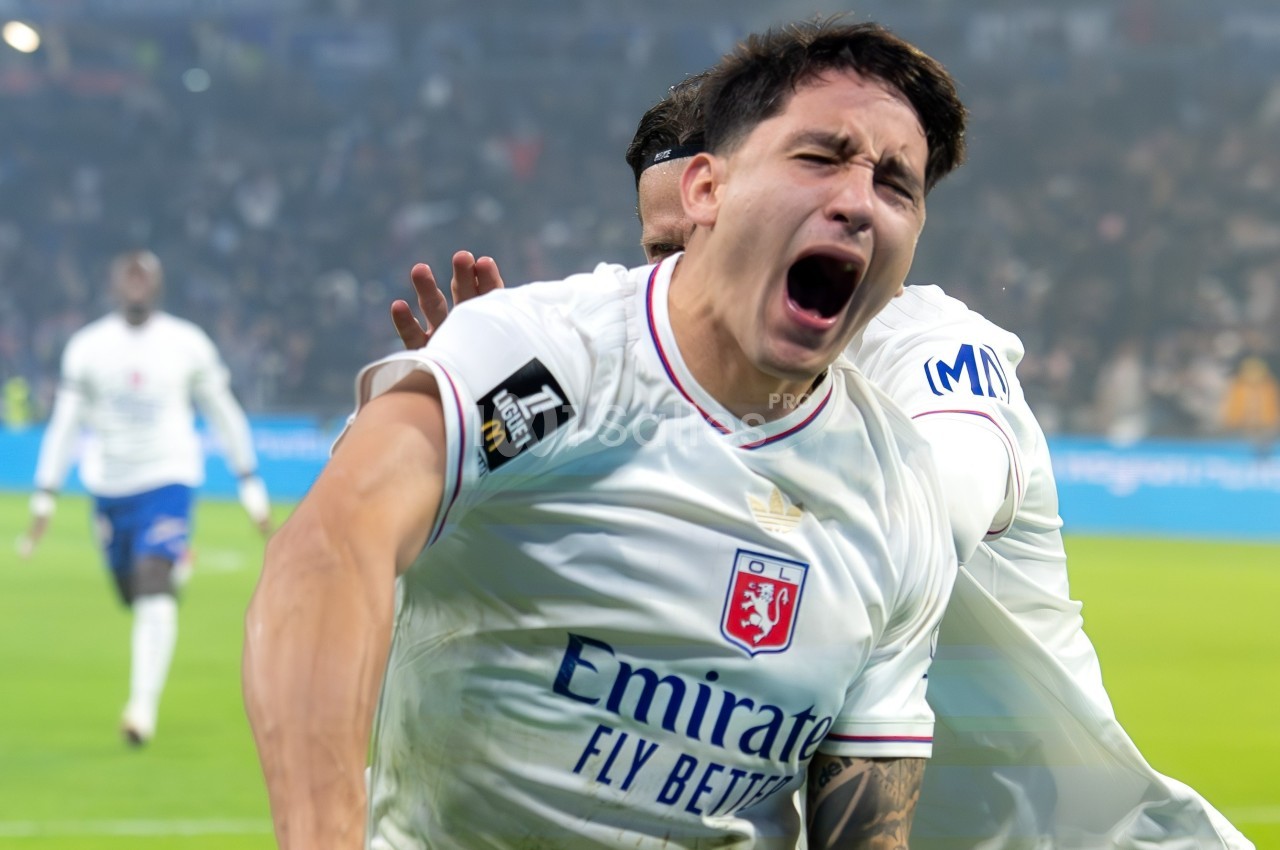 Un joueur de football en maillot blanc célèbre un but avec intensité sur un terrain devant un public flou.