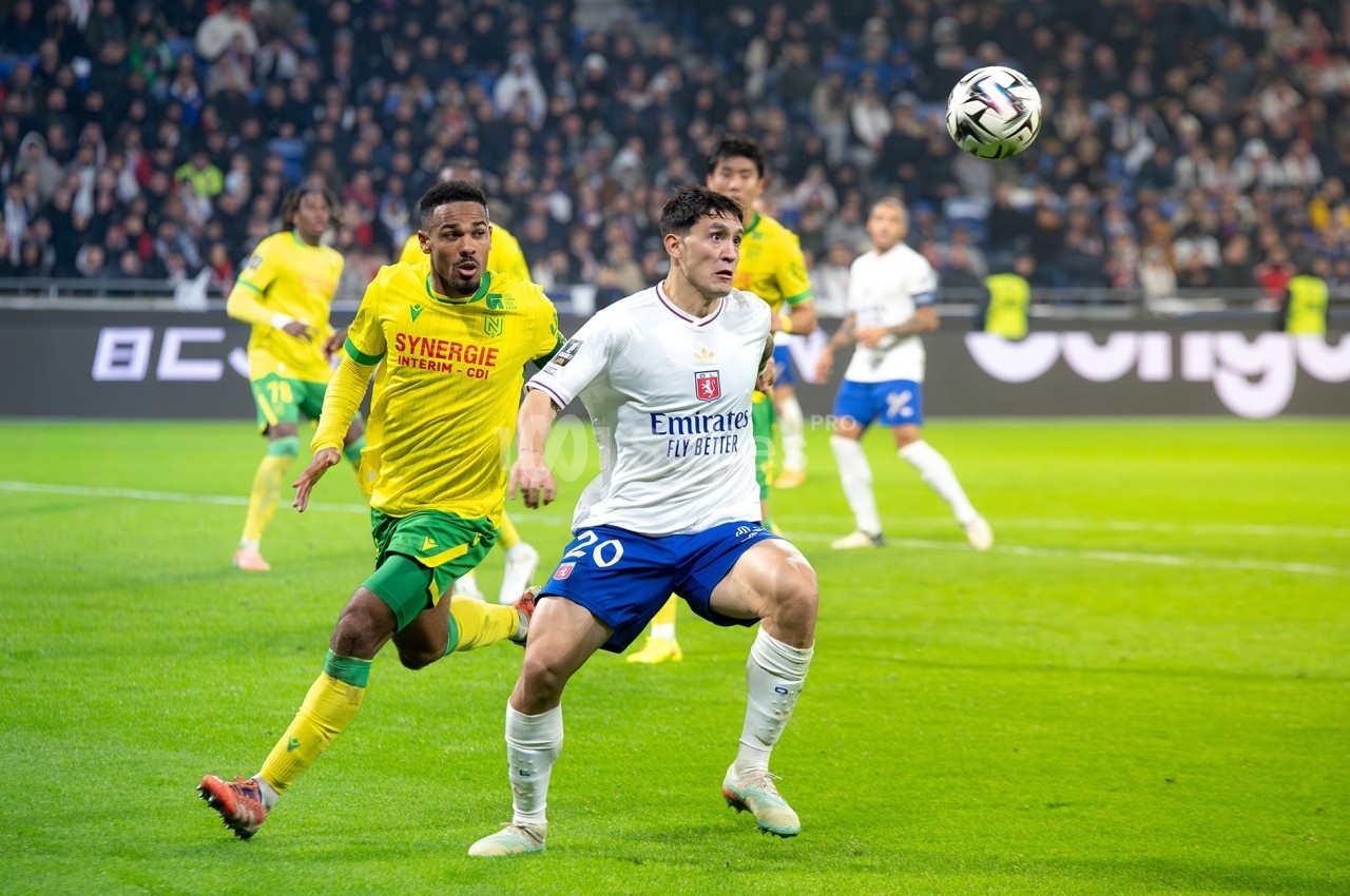 Un joueur de football en maillot blanc contrôle le ballon sous la pression d'un adversaire en maillot jaune sur un terrain.