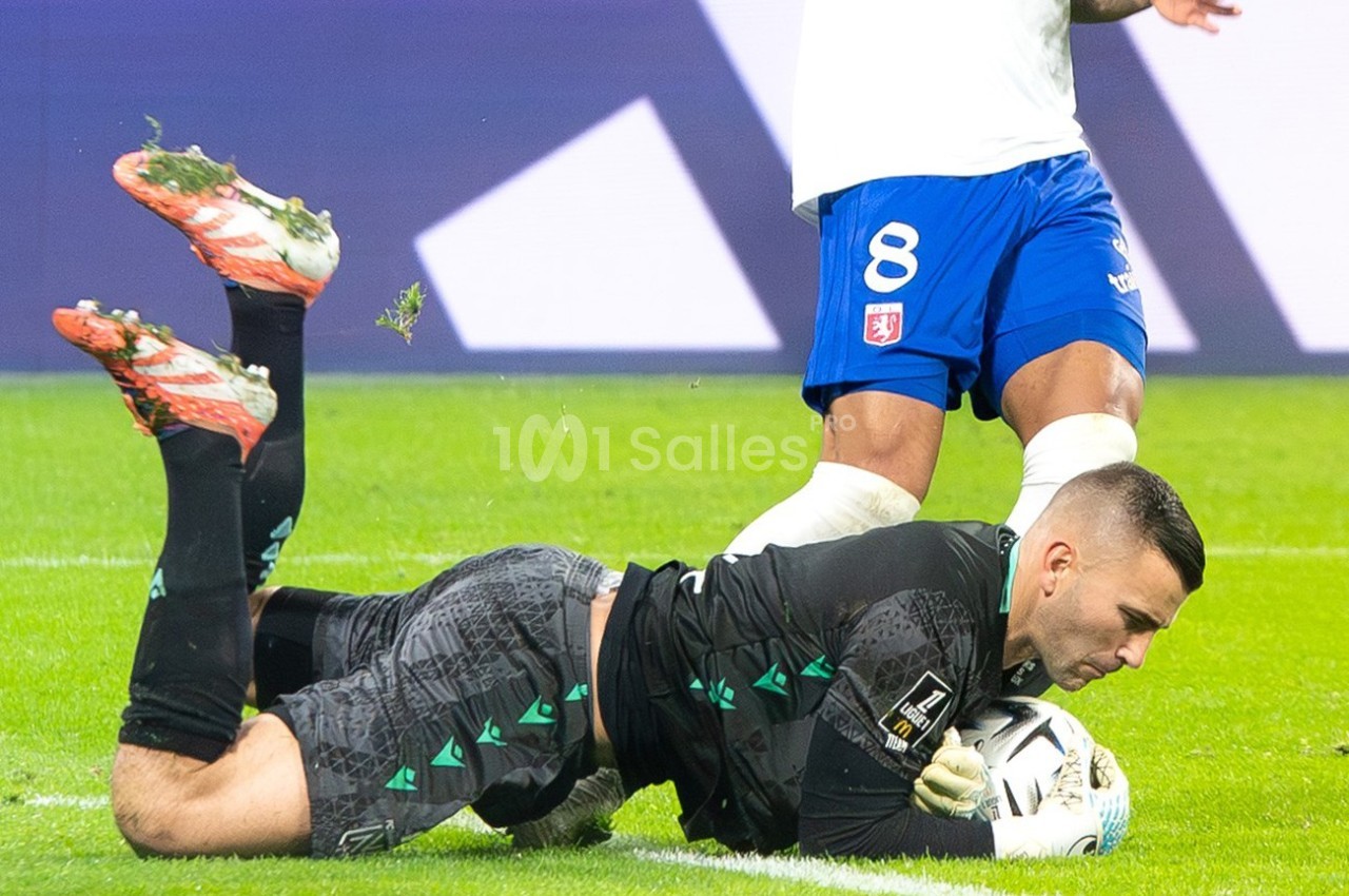 Un gardien de but en tenue noire attrape un ballon au sol lors d'un match de football, devant un joueur en blanc et bleu.