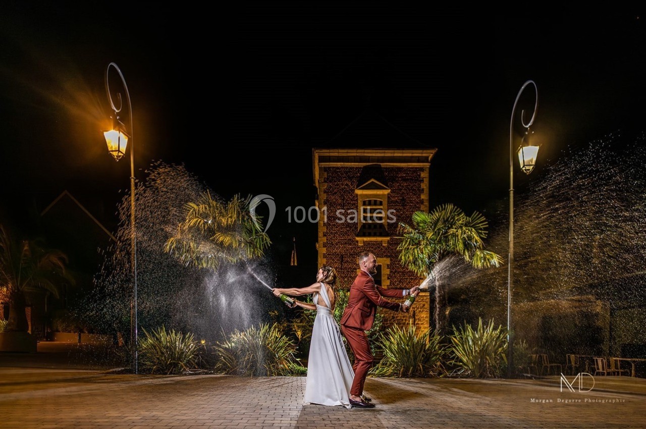 Un couple en tenue de mariage ouvre des bouteilles de champagne, créant des éclaboussures sous des lampadaires la nuit.