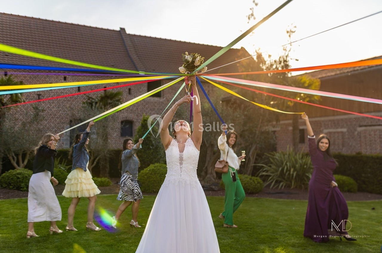 Une mariée en robe blanche tient un bouquet au centre de rubans colorés, entourée de femmes dans un jardin.
