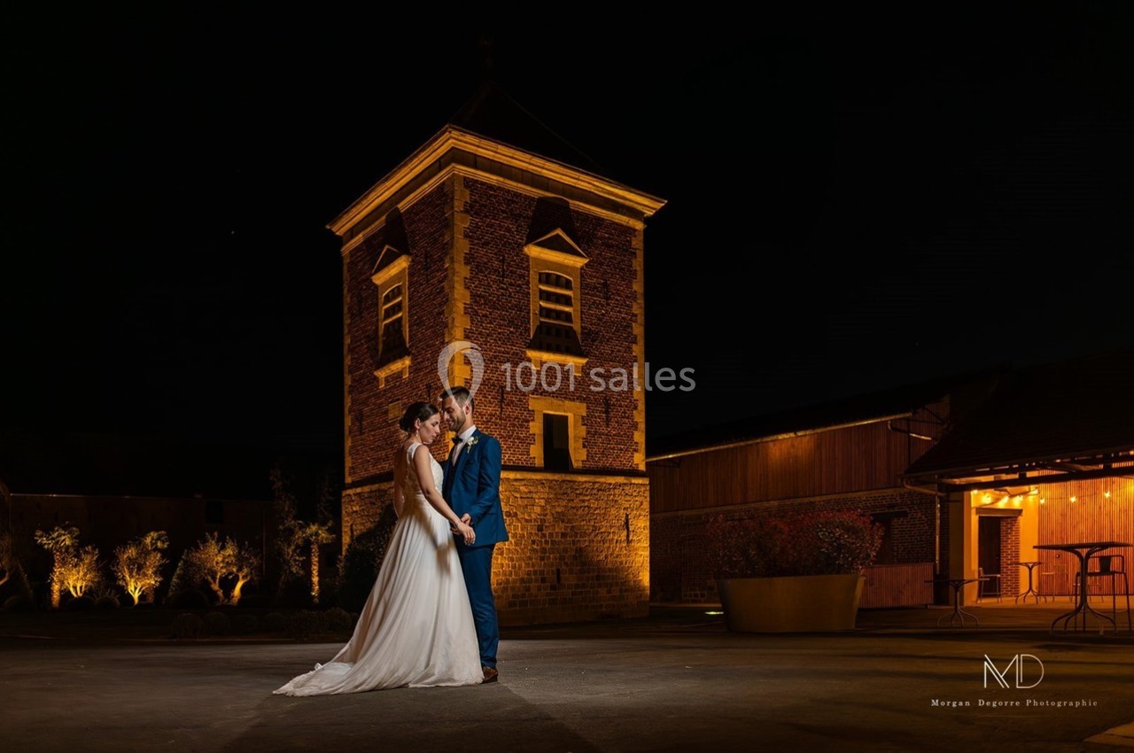 Un couple en tenue de mariage pose devant un bâtiment en briques éclairé la nuit.