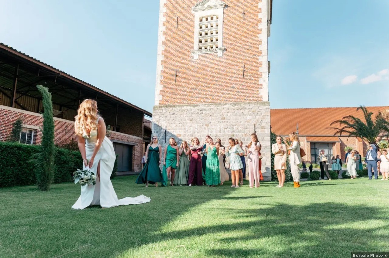 Une mariée en robe blanche s'apprête à lancer son bouquet devant un groupe d'invitées rassemblées sur une pelouse.