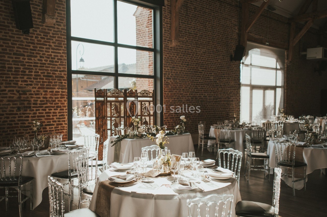 Salle de réception lumineuse avec murs en briques, grandes fenêtres et tables élégamment dressées pour un événement.