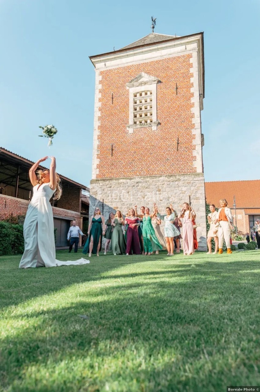 Une mariée lance son bouquet devant un groupe d'invités sur une pelouse, près d'un bâtiment en briques.