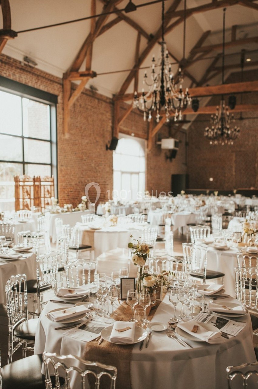 Salle de réception lumineuse avec tables rondes décorées de nappes blanches, chaises transparentes et chandeliers suspendus.
