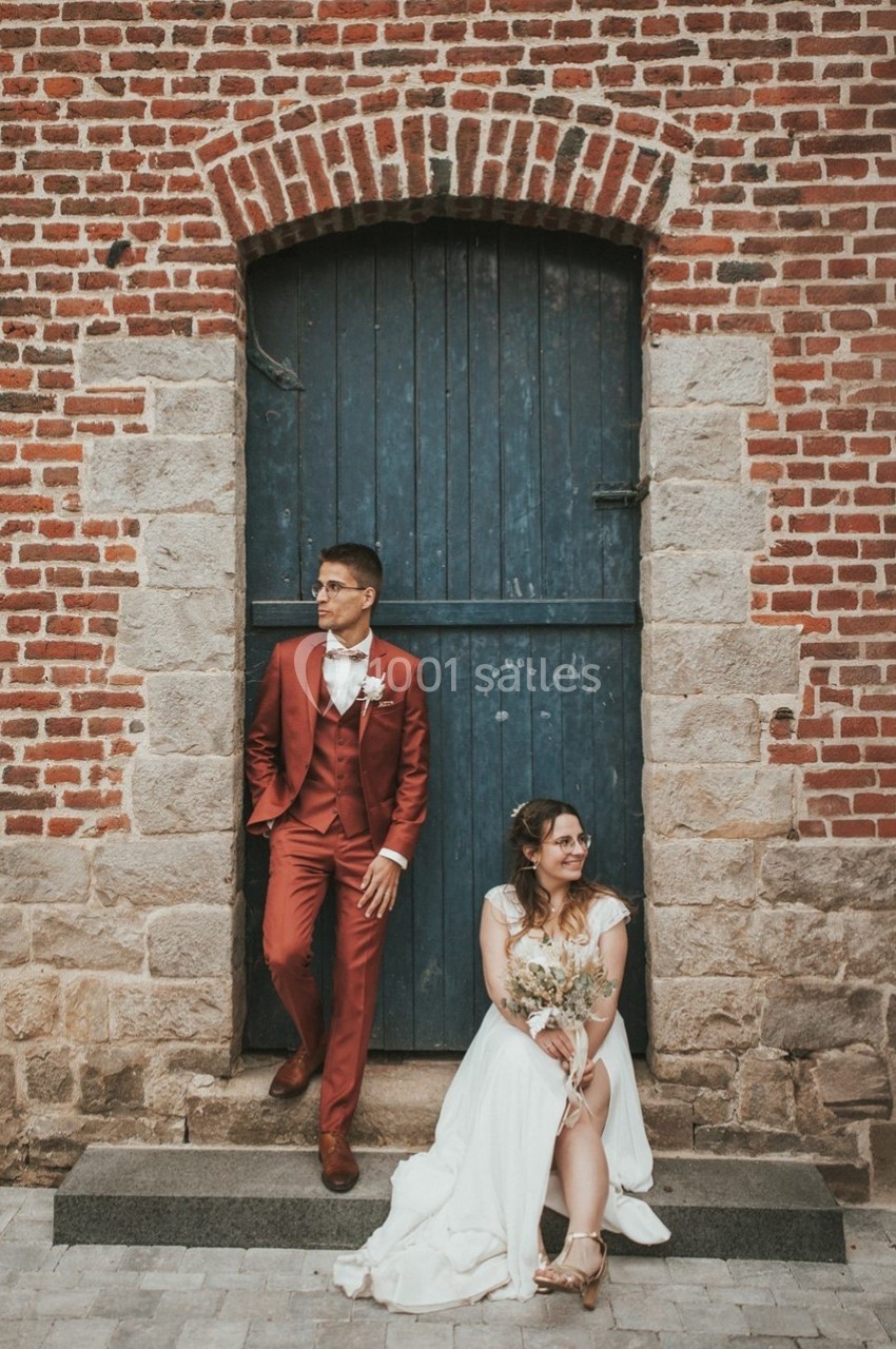 Un couple en tenue de mariage pose devant une porte bleue encadrée de briques rouges.
