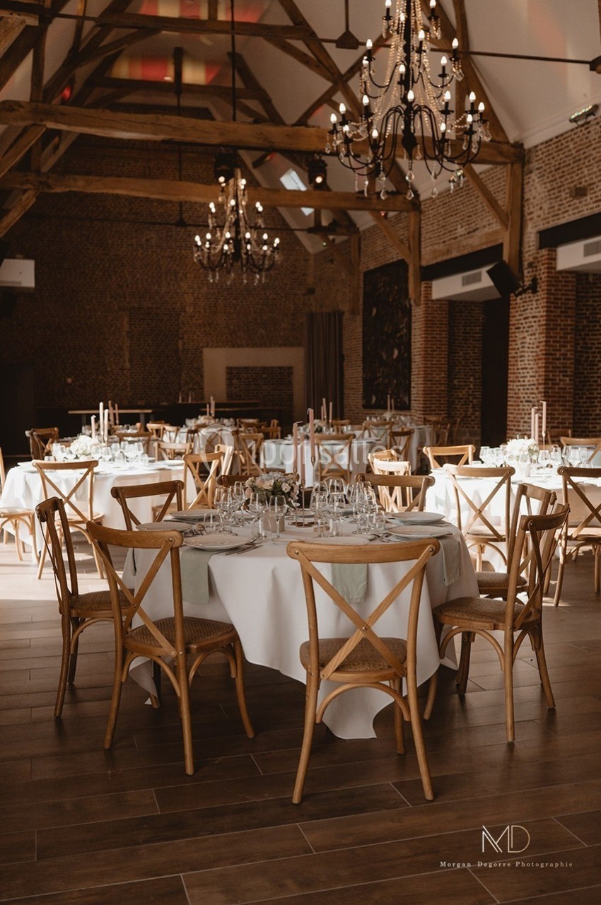 Salle de réception avec tables rondes dressées, chaises en bois, nappes blanches et lustres suspendus.