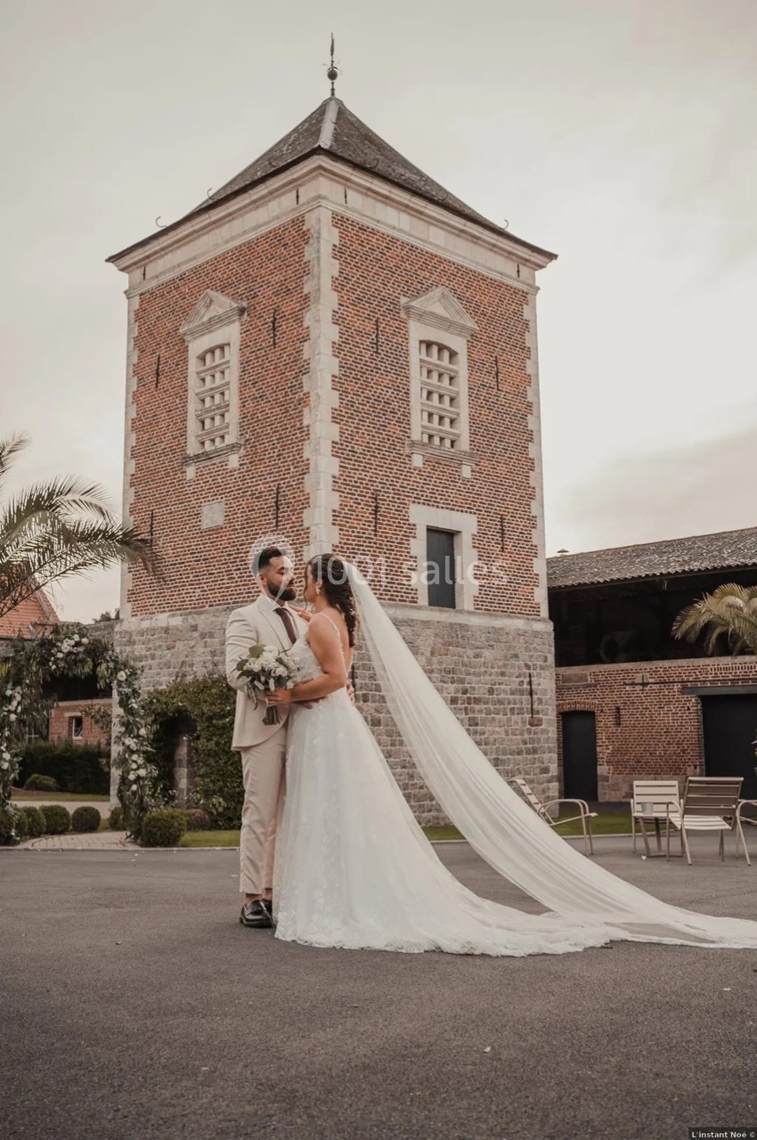 Un couple de mariés pose devant une tour en briques rouges dans une cour pavée, sous un ciel légèrement nuageux.