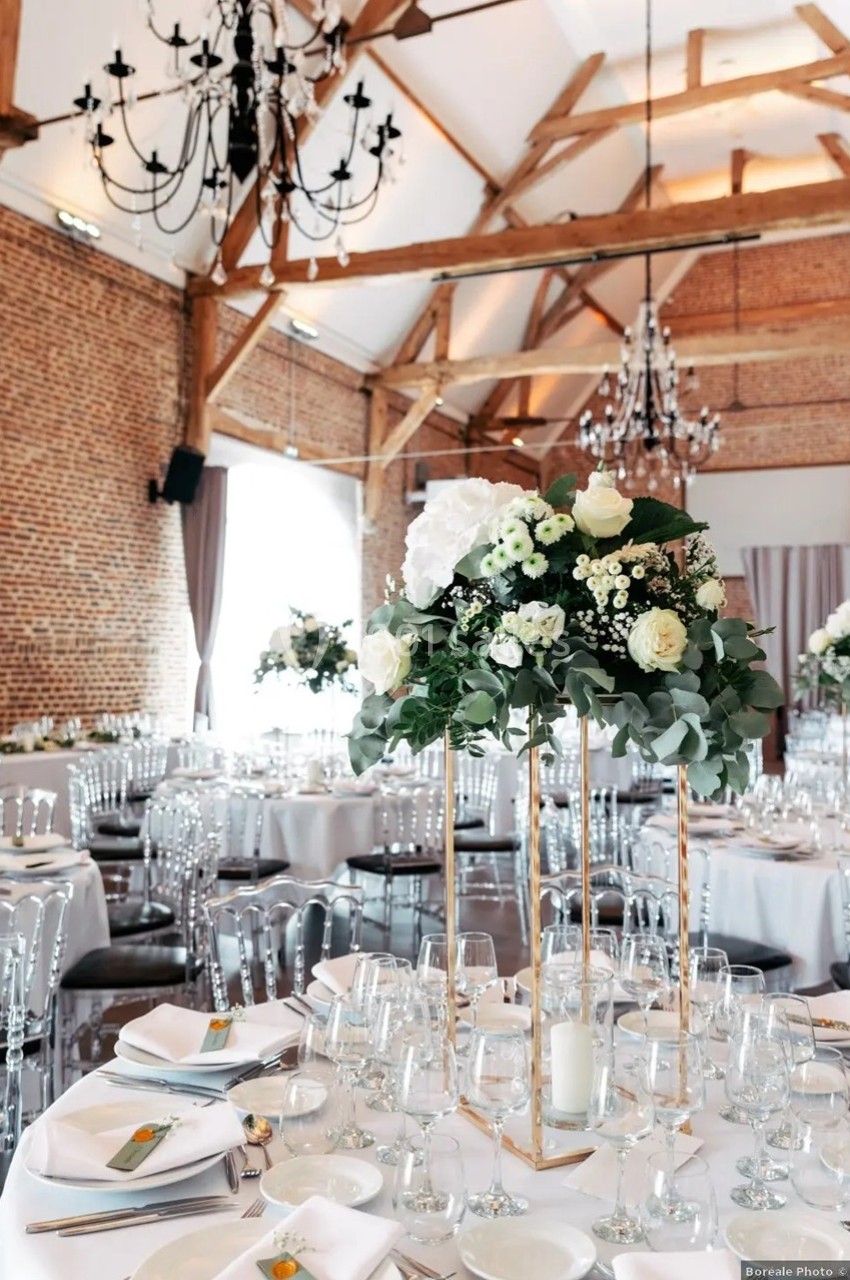 Salle de réception lumineuse avec tables décorées de centres de table floraux et poutres apparentes au plafond.