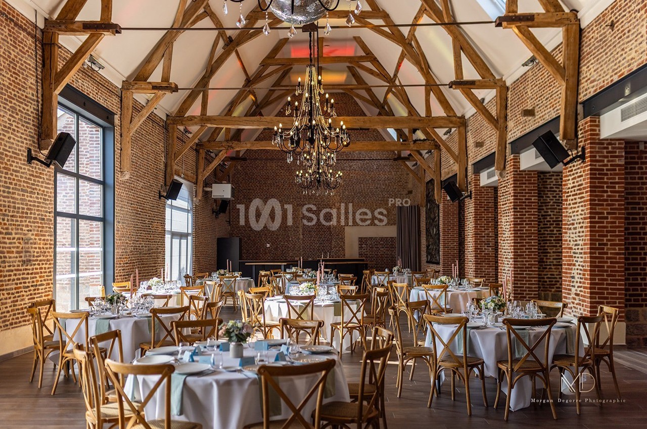 Salle de réception avec poutres apparentes, murs en briques, tables rondes dressées et lustre central élégant.